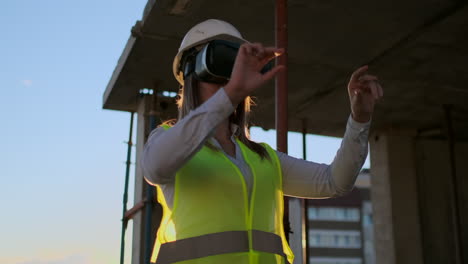 Woman-Engineer-Builder-on-the-roof-of-the-building-at-sunset-stands-in-VR-glasses-and-moves-his-hands-using-the-interface-of-the-future.-Futuristic-engineer-of-the-future.-The-view-from-the-back.